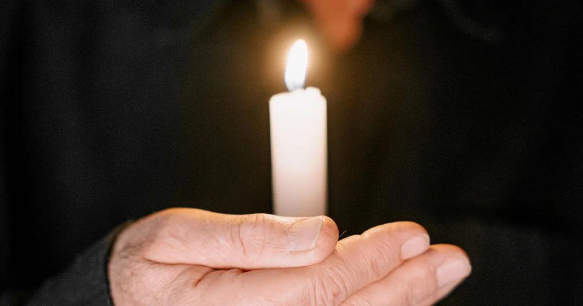A hand holding a lit white candle against a dark background — prayer and remembrance at the one-year memorial (pomana de un an)